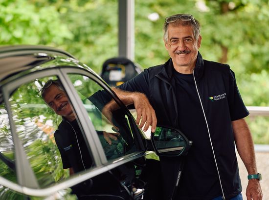 Ein Mann lächelt und steht neben einem Fahrzeug, während sein Spiegelbild im Seitenspiegel zu sehen ist. Er trägt eine schwarze Jacke mit Logo.