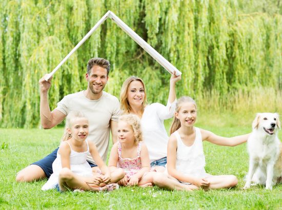 Eine Familie mit zwei Erwachsenen und drei Kindern sitzt auf einer Wiese zusammen mit einem Hund. Sie halten ein Holzgestell wie ein Hausmodell über sich.