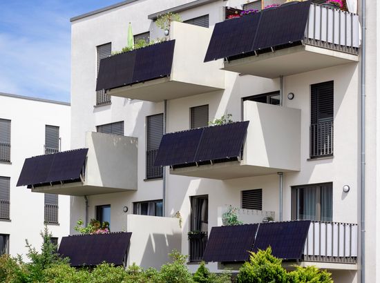 Moderne Wohngebäude mit Solarpanelen auf den Balkonen und begrünten Außenbereichen. Mehrstöckiger Bau mit großen Fenstern und geschwungenen Balkonstrukturen.