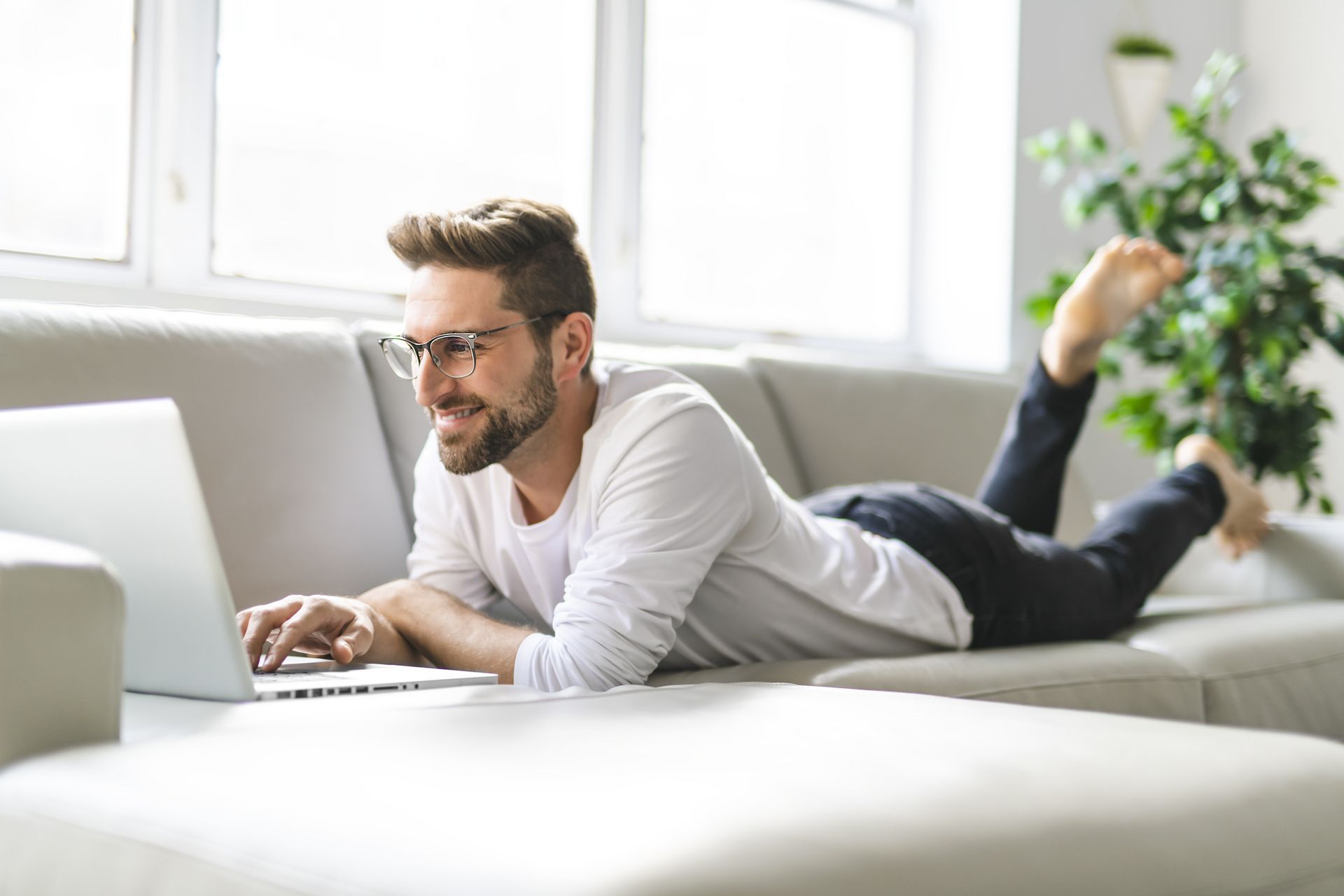 Ein Mann liegt entspannt auf einem Sofa und arbeitet auf einem Laptop, während er ein Bein hochgelegt hat.