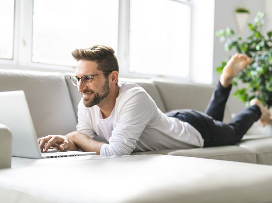 Ein Mann liegt entspannt auf einem Sofa und arbeitet auf einem Laptop, während er ein Bein hochgelegt hat.
