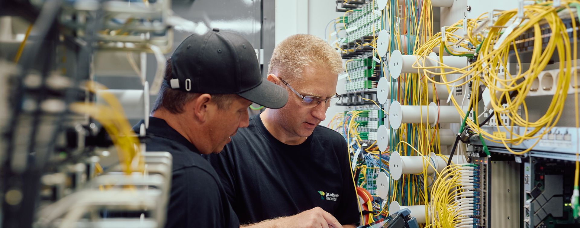 Zwei Techniker arbeiten an einer komplexen Netzwerk- oder Serverhardware mit vielen Kabeln und Anschlüssen. Sie führen Wartungs- oder Installationsarbeiten durch.