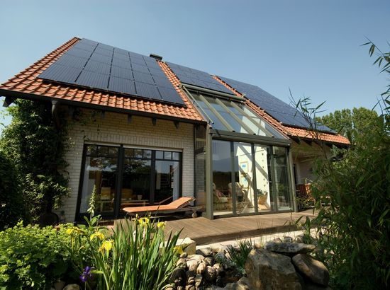 Moderne Hauserweiterung mit großem Wintergarten und Solarpanelen auf dem Dach, umgeben von üppiger Gartenbepflanzung.