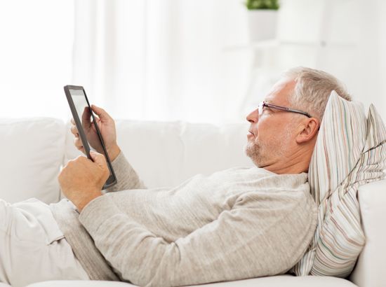 Ein älterer Mann liegt entspannt im Bett und hält ein Tablet in den Händen.