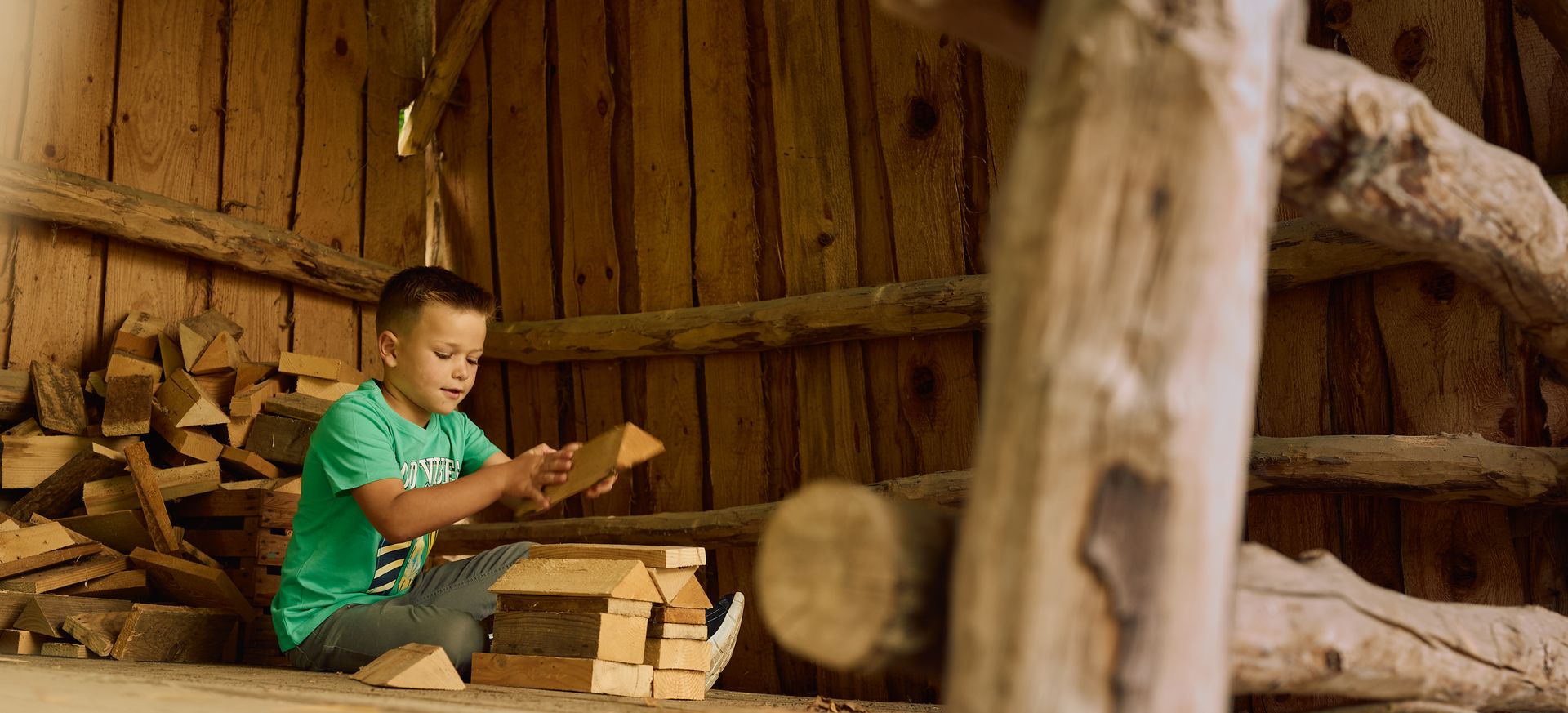 Ein Junge sitzt auf einer Holztreppe und hält ein Brett in den Händen, umgeben von geschnittenem Holz und Holzstapeln.