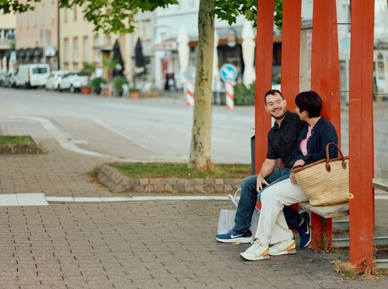Zwei Personen sitzen auf einer Bank und unterhalten sich auf einer belebten Straße in einer Stadt. Die Person links trägt eine Jacke und Jeans, die Person rechts eine Jacke und hält eine Einkaufstasche.