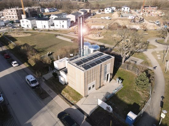 Luftaufnahme eines mehrstöckigen Gebäudes mit Solaranlage auf dem Dach in einer Wohnsiedlung mit angrenzendem Grünbereich und Parkplatz.