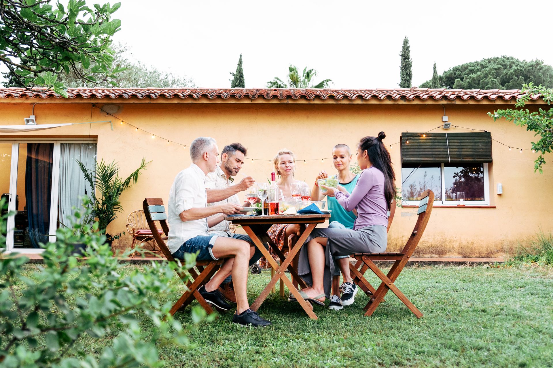 Eine Gruppe von vier Personen sitzt an einem Tisch im Freien und isst gemeinsam. Die Szene spielt in einem Garten vor einem gelben Haus.