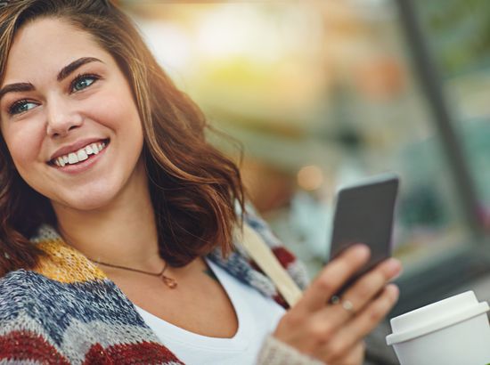 Eine lächelnde Person hält ein Smartphone in der Hand und scheint sich zu freuen, während sie einen Becher in der anderen Hand hält.