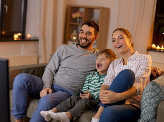 Eine Familie mit drei Personen sitzt auf einem Sofa und schaut gemeinsam lächelnd fern.