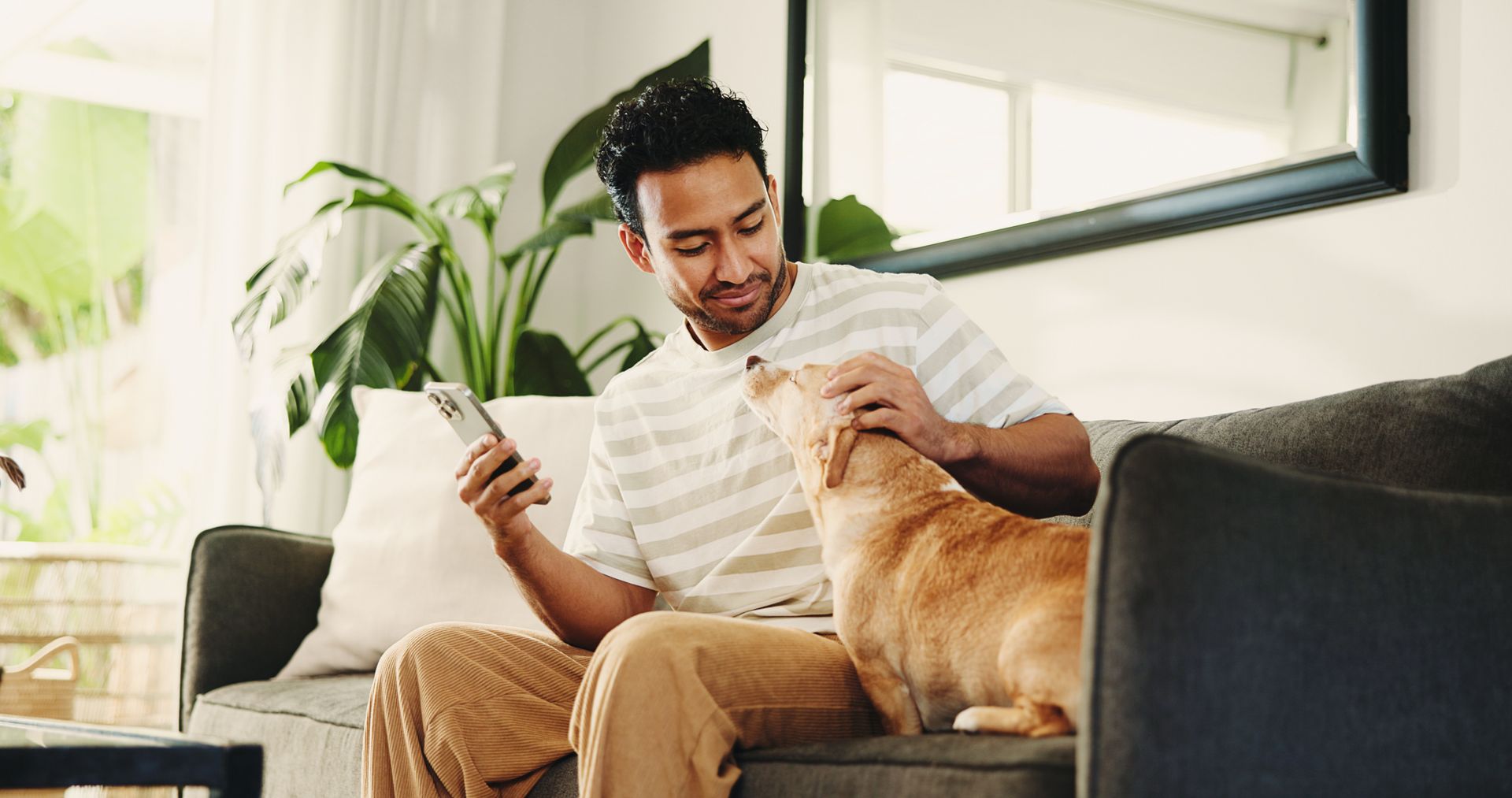 Ein Mann sitzt auf einem Sofa und hält ein Smartphone in der Hand, während er einen Hund streichelt.