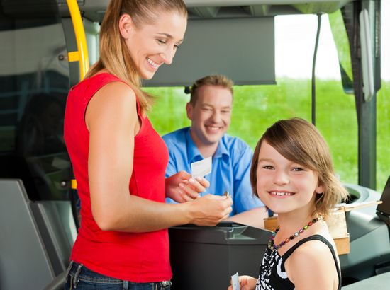 Eine Frau und ein Mädchen halten Fahrkarten in einem Bus, während ein Mann im Hintergrund lächelt. Die Szene spielt im Inneren eines öffentlichen Verkehrsmittels.