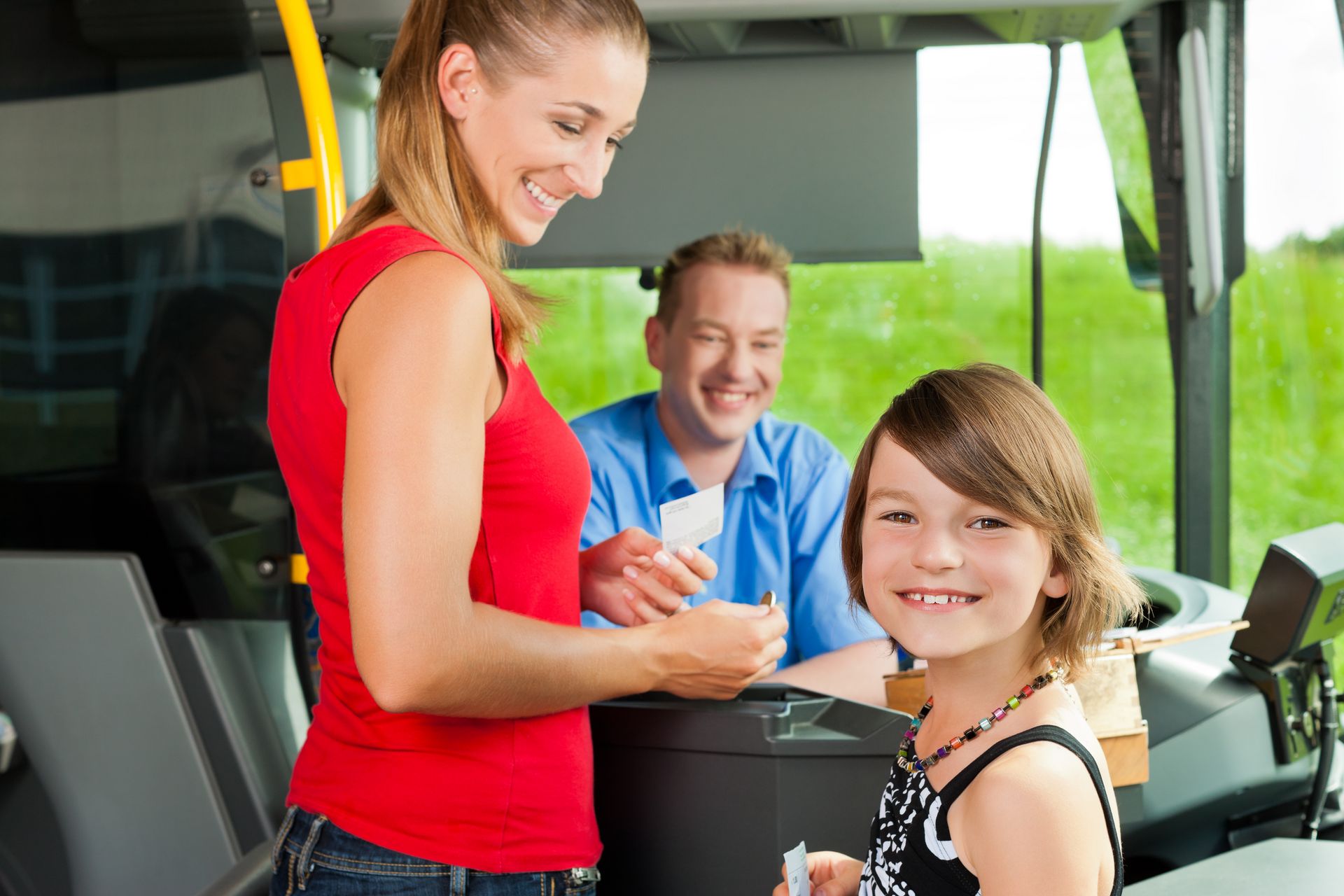 Eine Frau und ein Mädchen halten Fahrkarten in einem Bus, während ein Mann im Hintergrund lächelt. Die Szene spielt im Inneren eines öffentlichen Verkehrsmittels.
