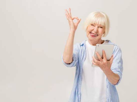 Eine ältere Frau hält ein Tablet und zeigt mit der anderen Hand ein Daumen-Okay-Zeichen, während sie lächelnd nach oben schaut.