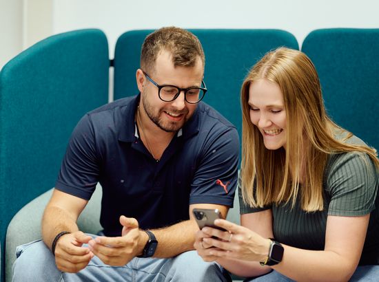 Zwei Personen sitzen auf einem Sofa und schauen gemeinsam auf ein Smartphone, wobei sie lächelnd und konzentriert wirken.