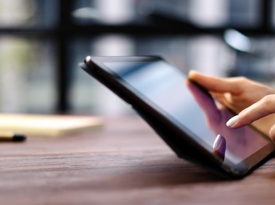 Person nutzt ein Tablet mit digitalen Notizen oder einer Anwendung am Tisch, umgeben von Büchern und Stiften.
