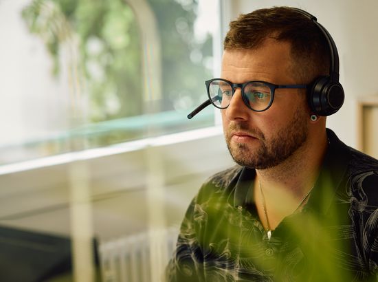Ein Mann mit Kopfhörern und Brille sitzt konzentriert und blickt nachdenklich zur Seite. Er trägt ein grünes Oberteil.
