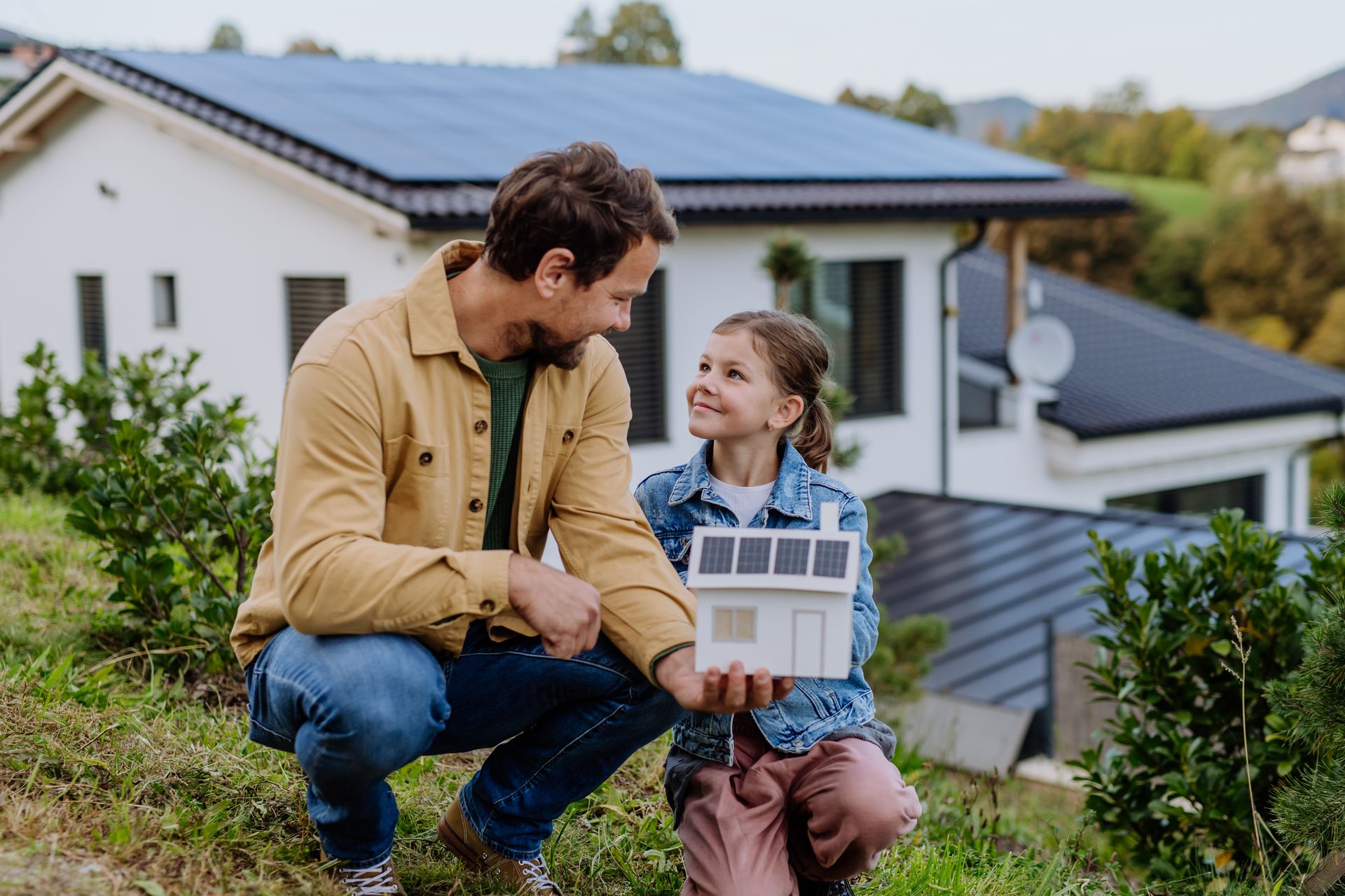 Ein Mann und ein Kind sitzen im Garten und betrachten gemeinsam ein Modellhaus mit Solaranlage auf dem Dach.