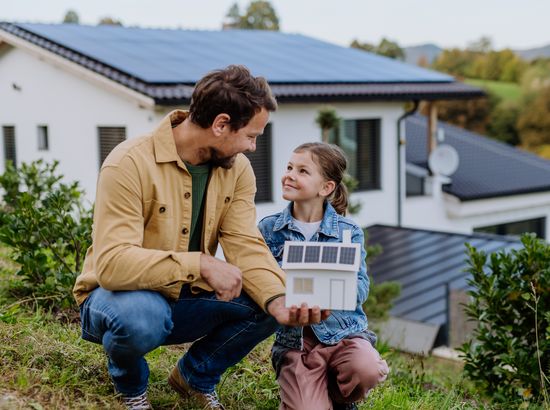 Ein Mann und ein Kind sitzen im Garten und betrachten gemeinsam ein Modellhaus mit Solaranlage auf dem Dach.