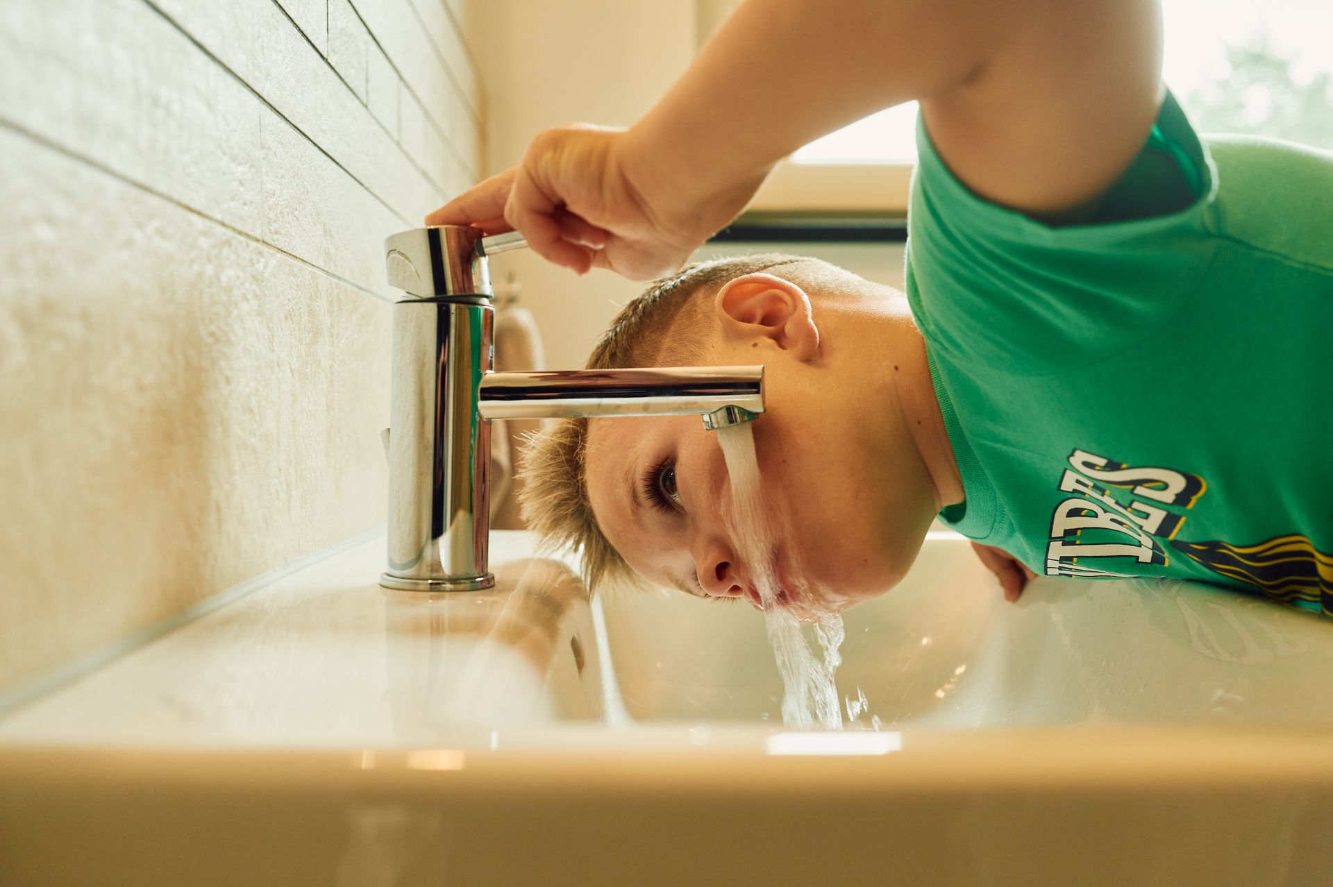 Ein Kind wäscht sich mit Wasser aus dem Hahn das Gesicht im Waschbecken.