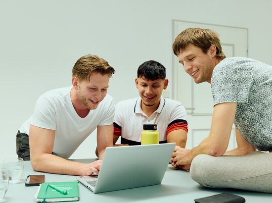 Drei Personen sitzen entspannt und schauen gemeinsam auf einen Laptop, der auf einem Tisch steht.