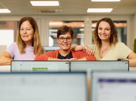 Drei Personen sitzen vor Computern in einem Büro und lächeln in die Kamera. Sie arbeiten gemeinsam an ihren Arbeitsplätzen.