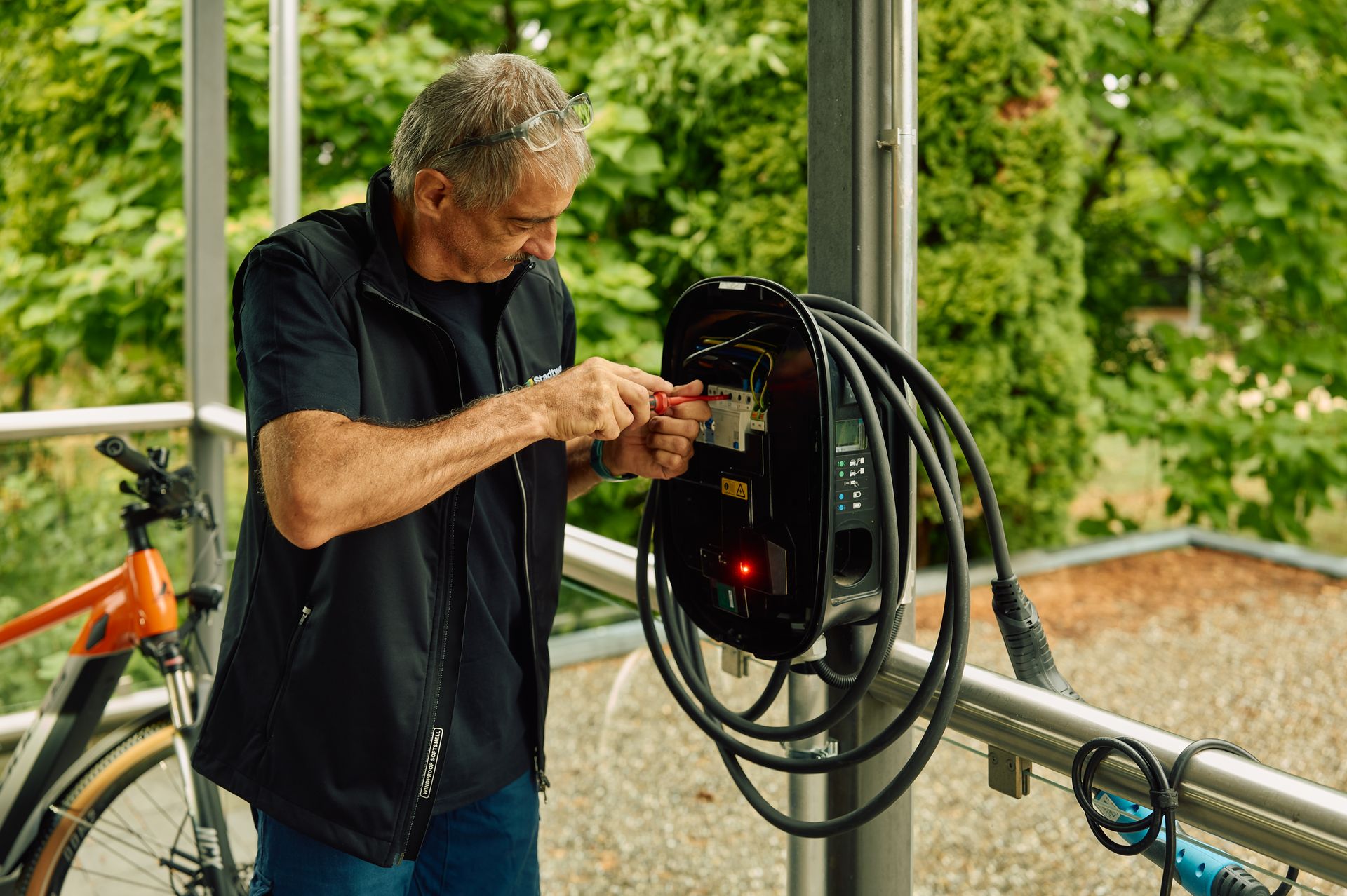 Ein Mann repariert oder justiert eine elektrische Ladestation für ein Fahrrad. Er hält ein Werkzeug in der Hand und arbeitet an der Anschlussbuchse.
