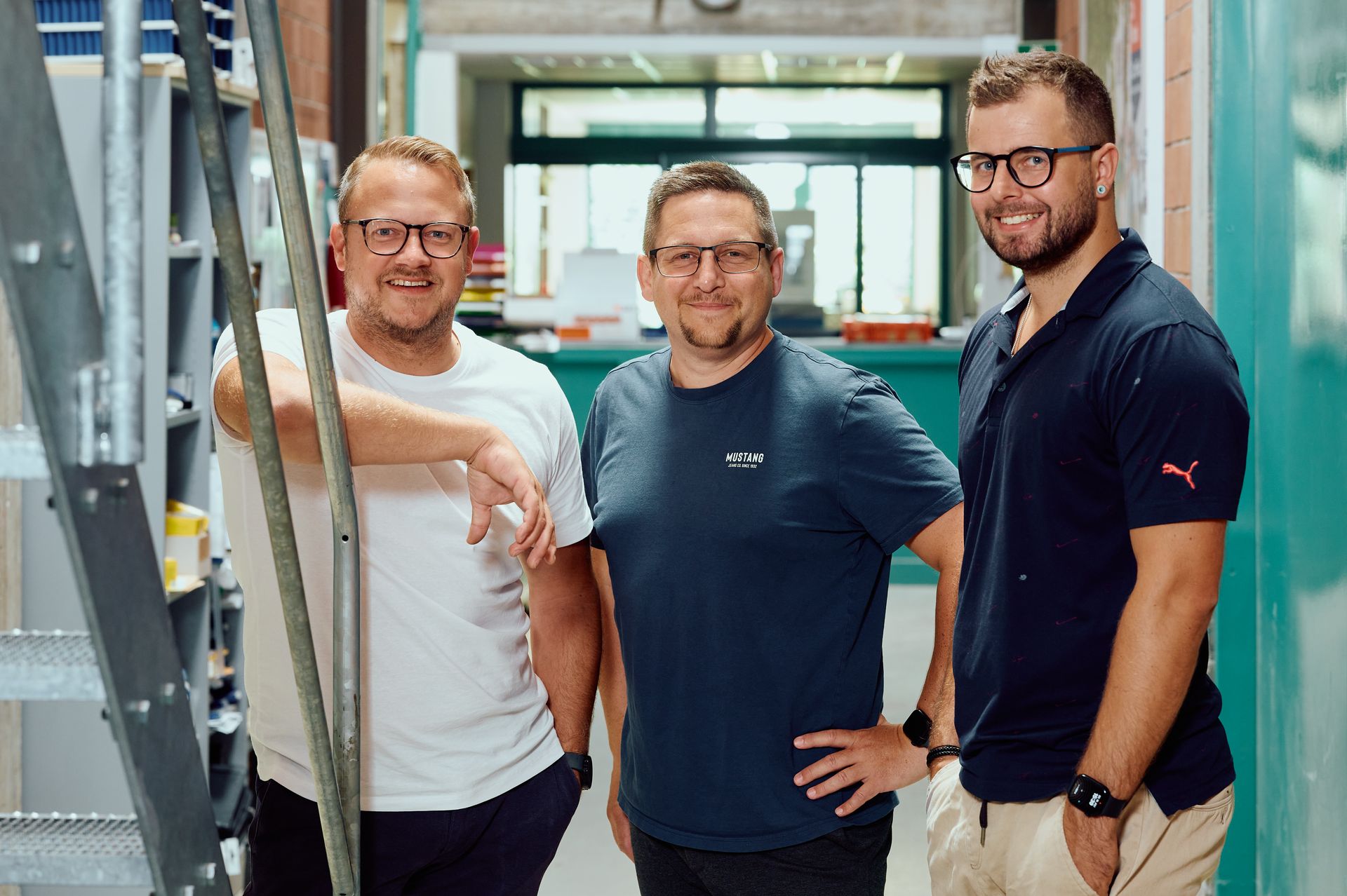 Drei Männer stehen lächelnd in einem Innenraum mit Industrieelementen im Hintergrund und posieren für ein Gruppenfoto.
