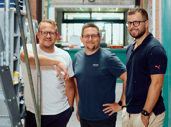 Drei Männer stehen lächelnd in einem Innenraum mit Industrieelementen im Hintergrund und posieren für ein Gruppenfoto.
