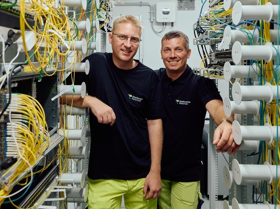Zwei Mitarbeiter stehen vor einem komplexen Kabel- und Verteilerregal in einem Technikraum und lächeln in die Kamera.