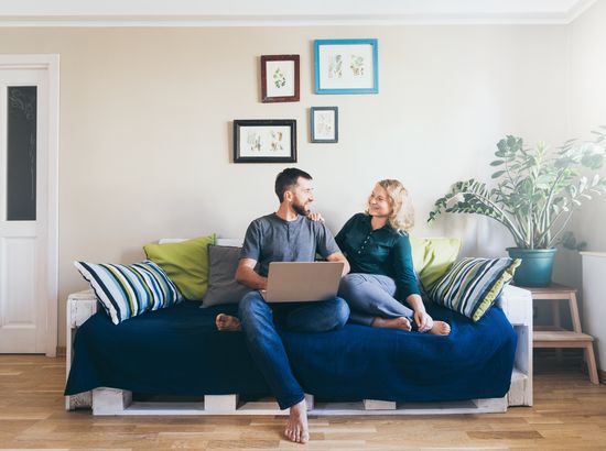 Zwei Personen sitzen auf einem Sofa und arbeiten gemeinsam an einem Laptop, während sie sich unterhalten.