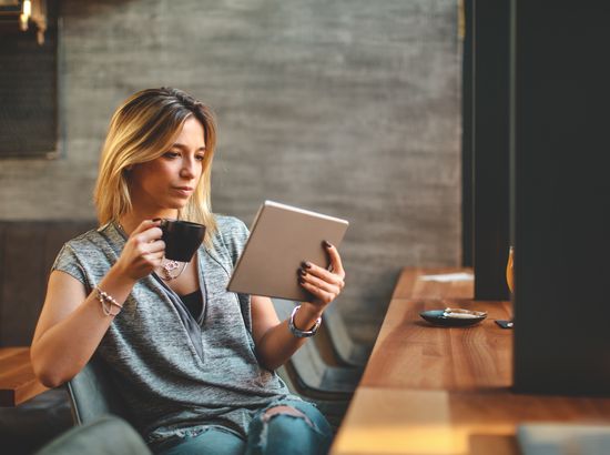 Eine Person sitzt in einem Café und hält ein Tablet in der einen sowie eine Tasse in der anderen Hand. Sie wirkt konzentriert und entspannt.
