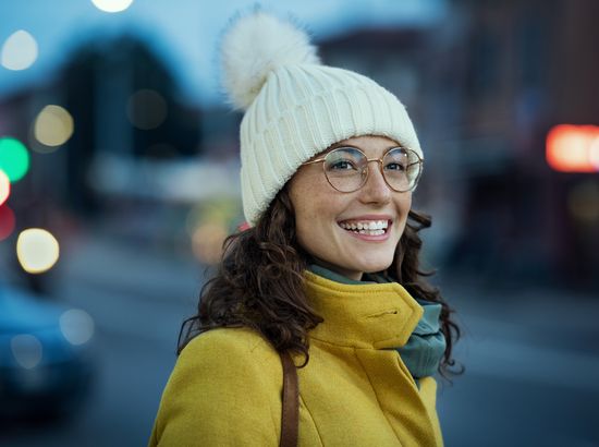 Eine lächelnde Person mit Brille und weißer Mütze trägt einen gelben Mantel und einen grünen Schal. Sie wirkt fröhlich und steht vor einem leicht unscharfen Hintergrund mit Lichtern.