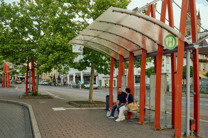 Zwei Personen sitzen auf einer Bank an einer Bushaltestelle mit rot-orangefarbenen Stützen und Dach. Im Hintergrund ist eine Straße mit Bäumen und Gebäuden zu sehen.