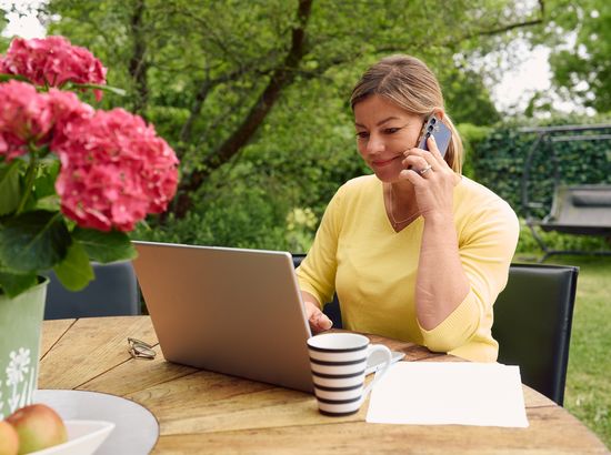 Eine Frau sitzt an einem Tisch im Freien und telefoniert, während sie auf einem Laptop arbeitet. Vor ihr stehen eine Tasse und ein Apfel.