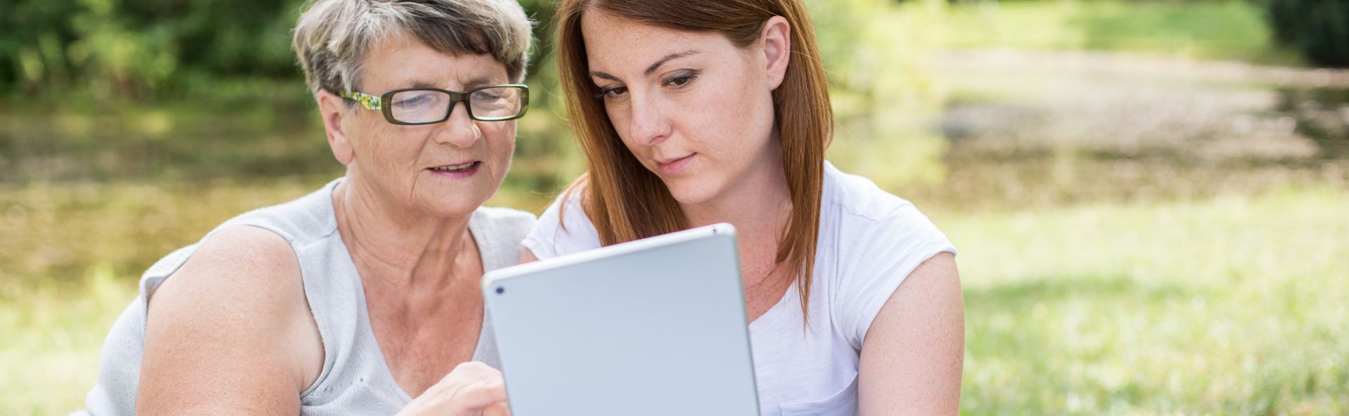 Zwei Personen schauen gemeinsam auf einen Tabletcomputer, während sie im Freien sitzen. Eine ältere Frau und eine jüngere Frau betrachten den Bildschirm aufmerksam.