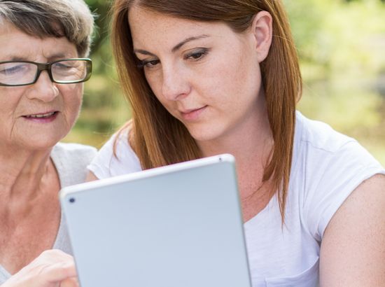Zwei Personen schauen gemeinsam auf einen Tabletcomputer, während sie im Freien sitzen. Eine ältere Frau und eine jüngere Frau betrachten den Bildschirm aufmerksam.