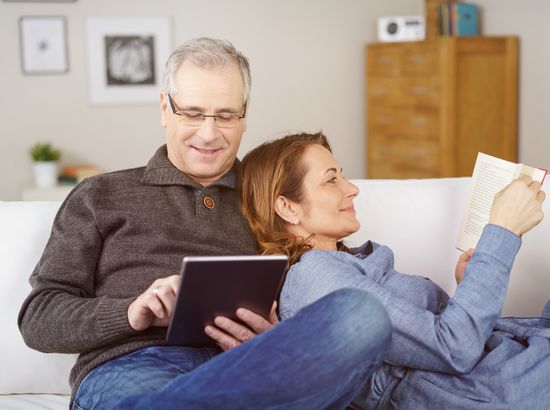 Ein älterer Mann und eine Frau sitzen auf einem Sofa, schauen gemeinsam auf ein Tablet und ein Buch. Beide wirken entspannt und beschäftigen sich mit digitalen und gedruckten Inhalten.