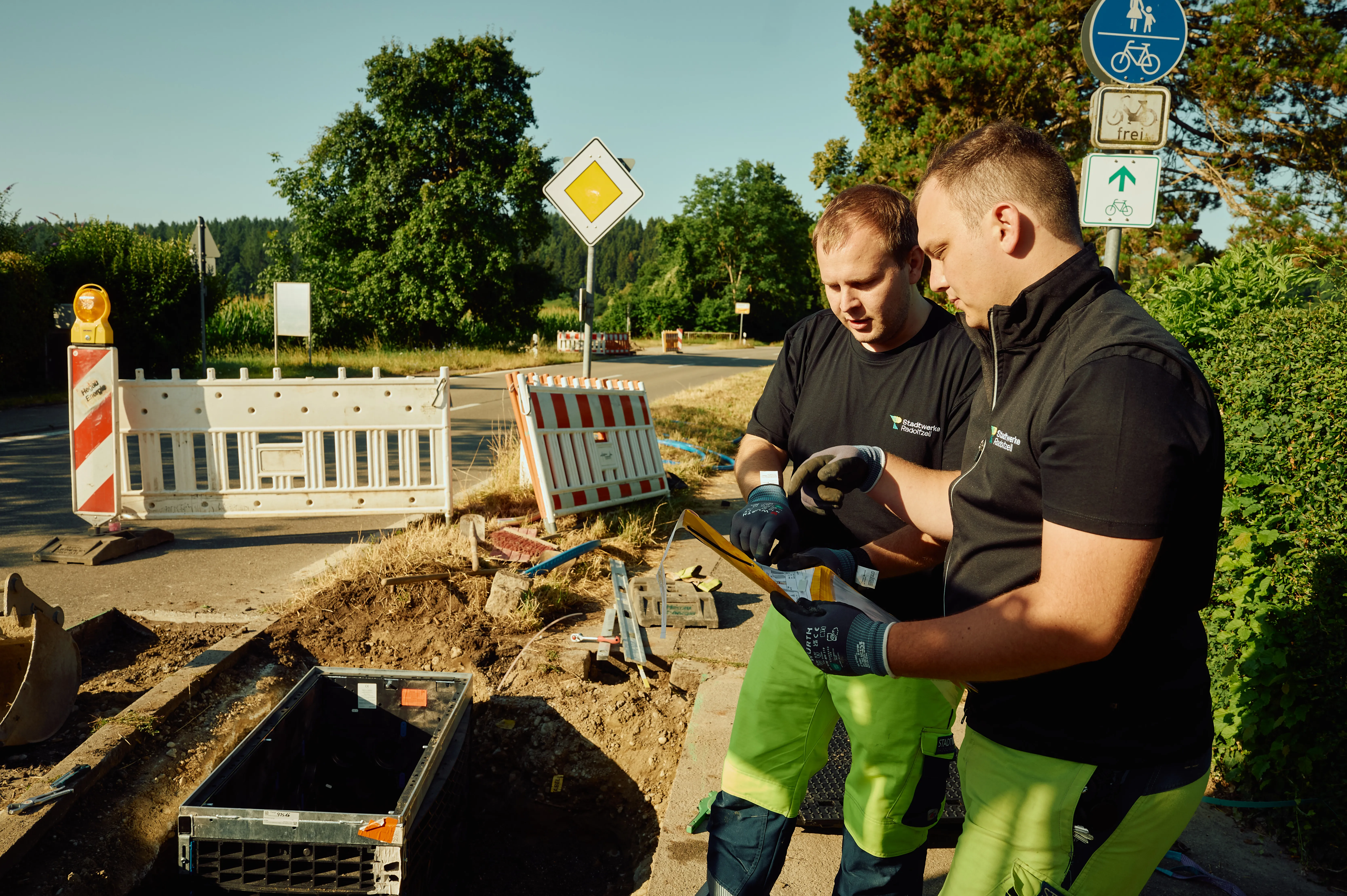 Zwei Arbeiter besprechen an einer Baustelle am Straßenrand die Installation von Infrastrukturkomponenten, während sie Schutzausrüstung tragen.