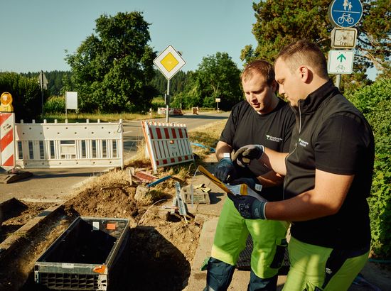 Zwei Arbeiter besprechen an einer Baustelle am Straßenrand die Installation von Infrastrukturkomponenten, während sie Schutzausrüstung tragen.