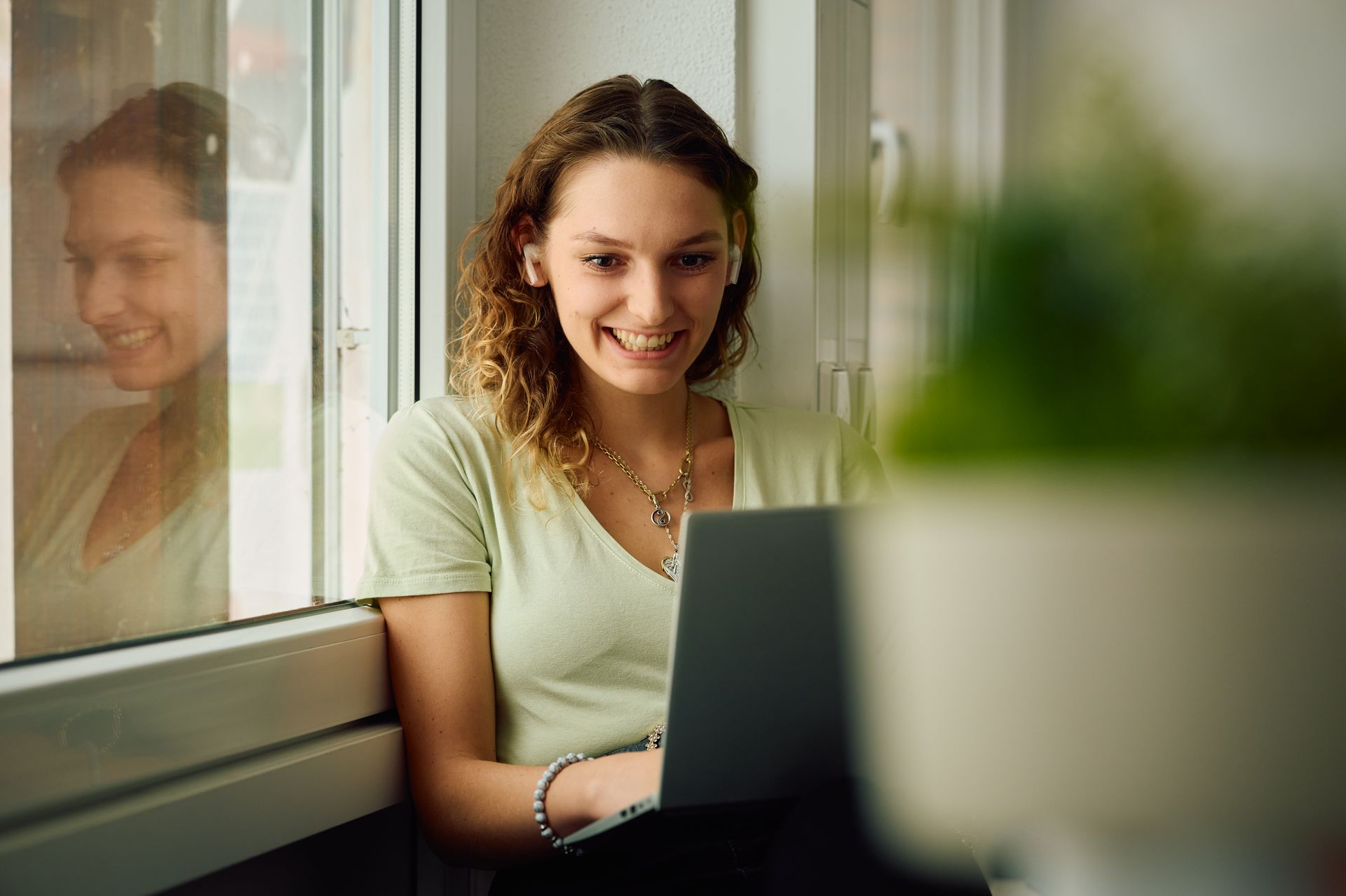 Eine Person mit Lächeln arbeitet an einem Laptop am Fenster sitzend. Sie trägt ein helles Oberteil und eine Kette.